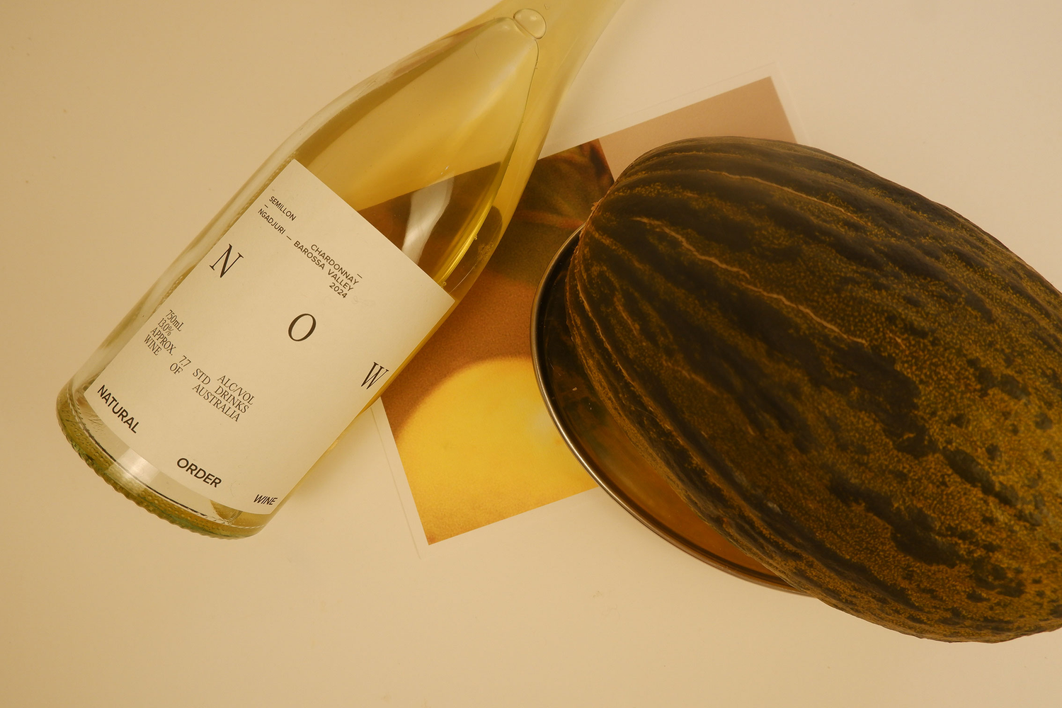 Bottle of white wine labeled 'NOW' next to a cantaloupe on a beige surface
