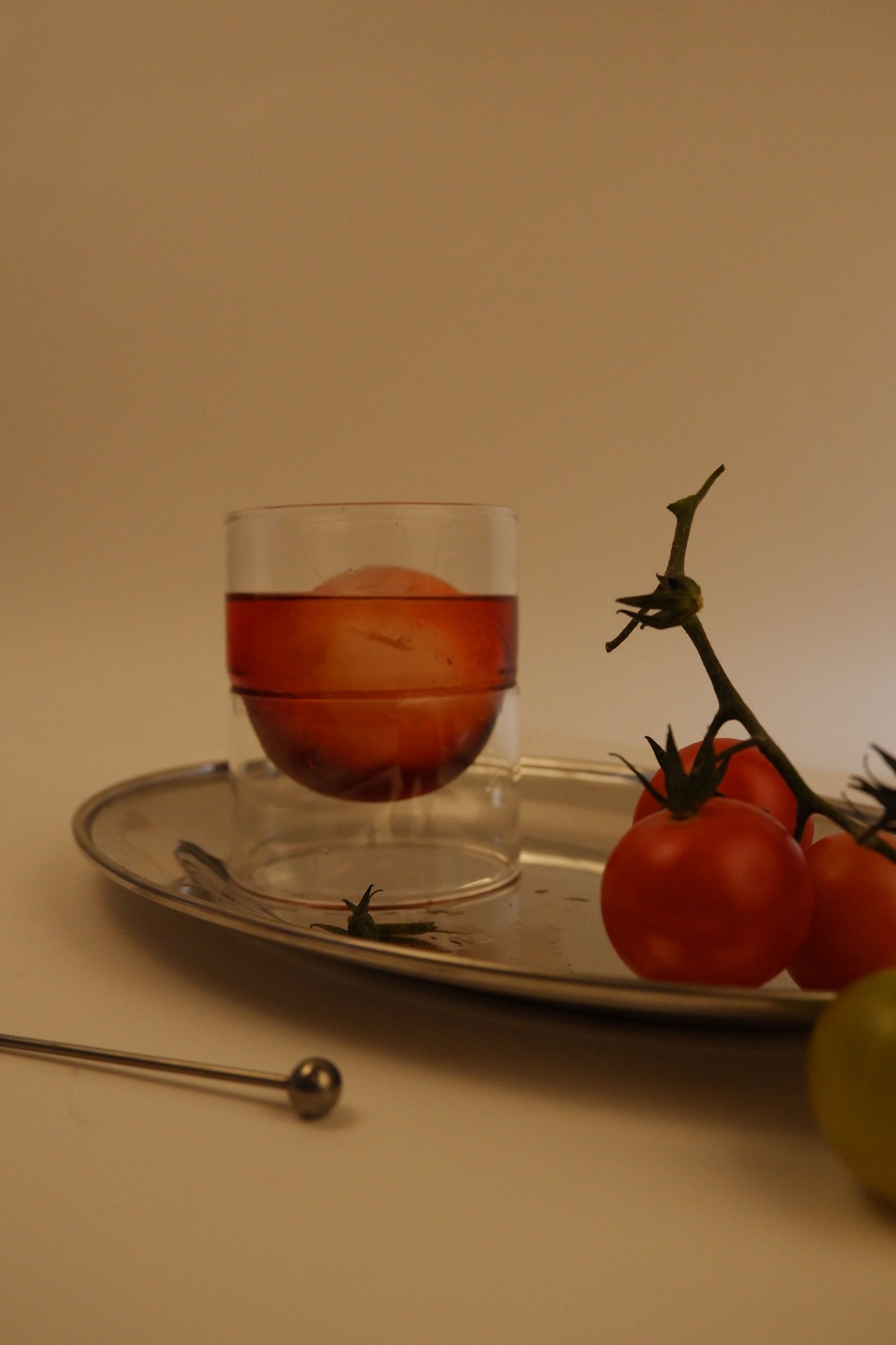 Glass with red liquid and fruit on a tray with tomatoes and a stick in the foreground.