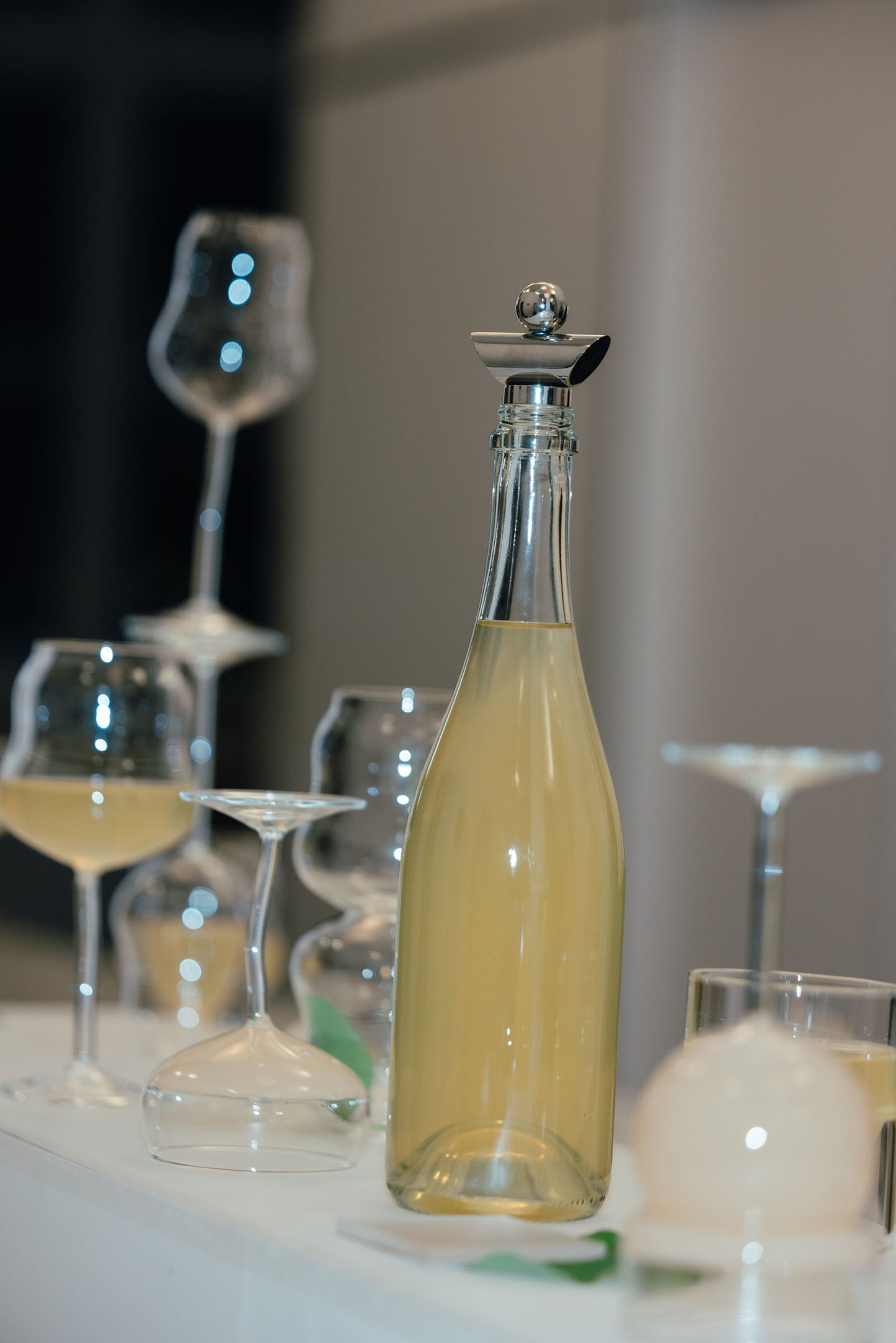 A clear glass bottle filled with pale yellow liquid, topped with a silver stopper, standing on a white surface with wine glasses and other glassware blurred in the background.