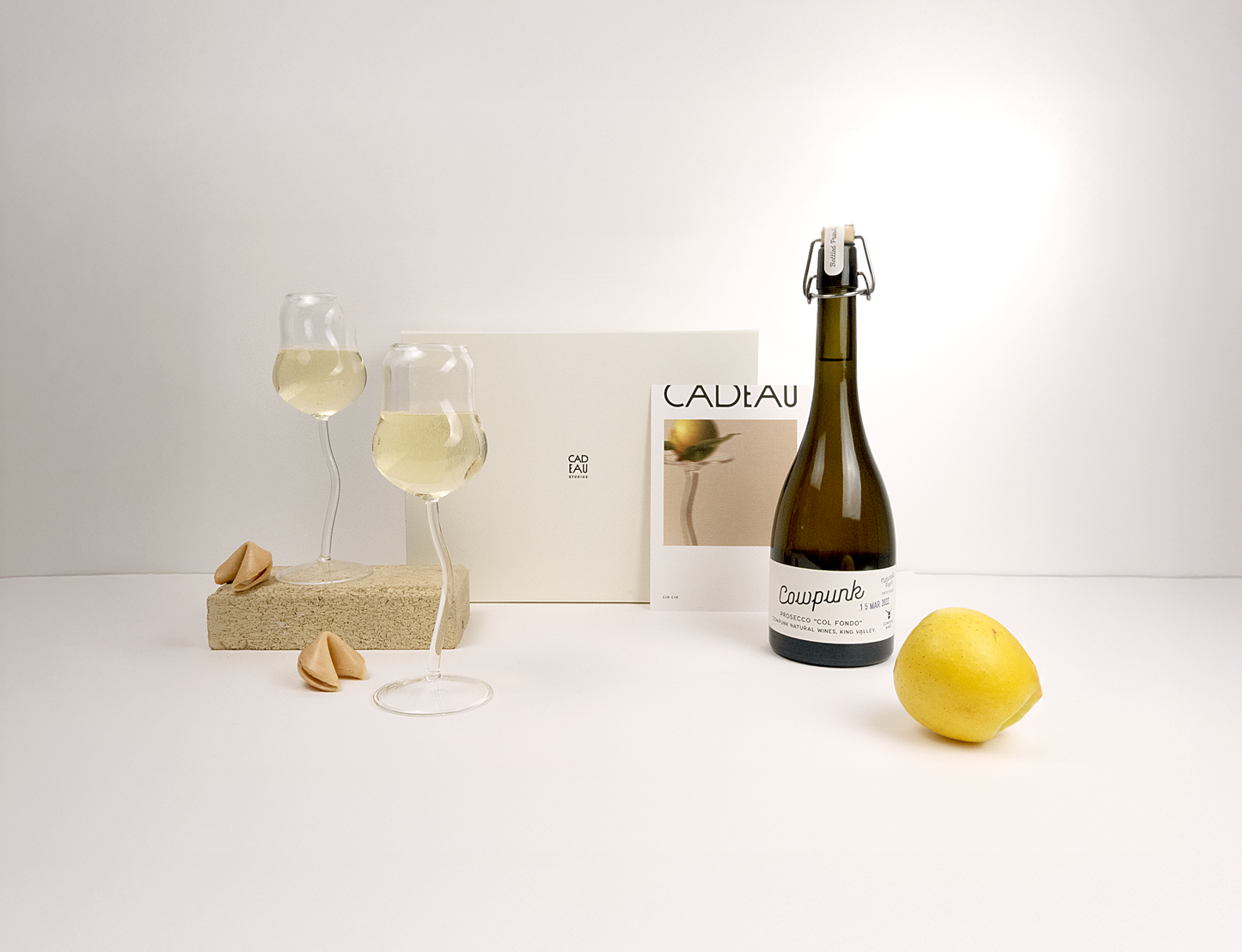 Bottle of white wine, two glasses with wine, a lemon, and a magazine on a white surface.
