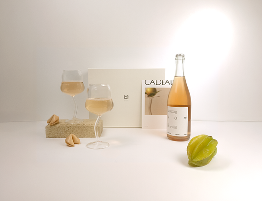 Bottle of Cadeau sparkling wine with two glasses and a star fruit on a white surface.