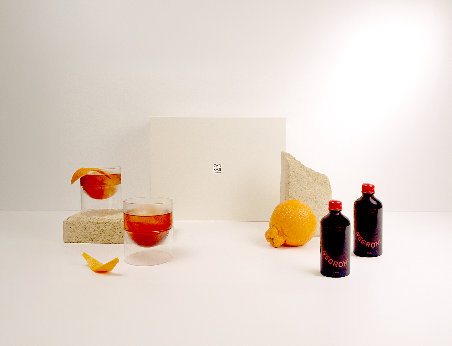 Glass of red liquid with a slice of fruit, lemon, and two bottles on a white surface.