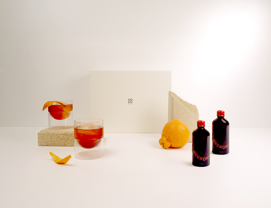 Glass of red liquid with a slice of fruit, lemon, and two bottles on a white surface.
