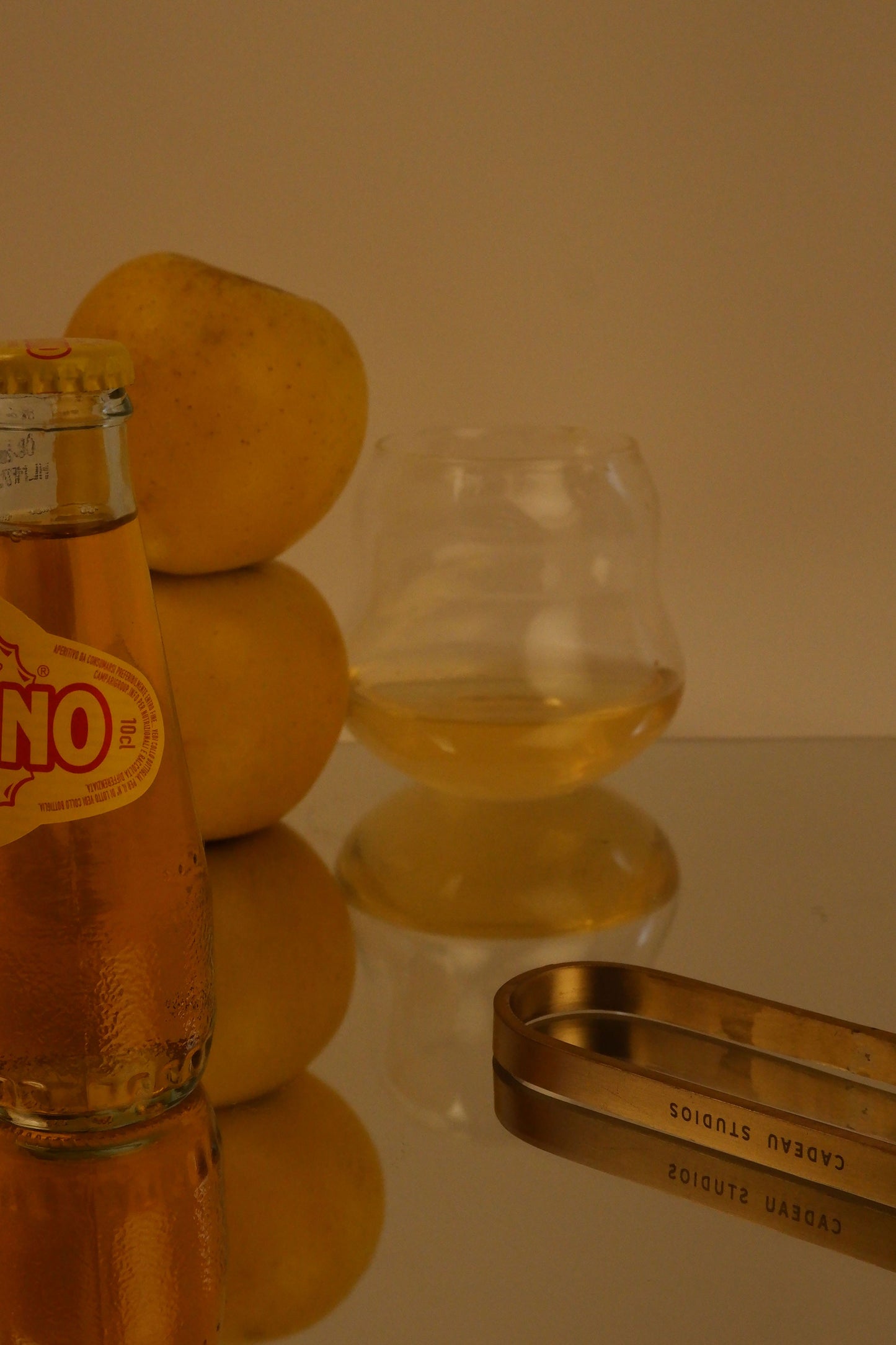 Lemon bottle, glass with liquid, and citrus fruit on a reflective surface.