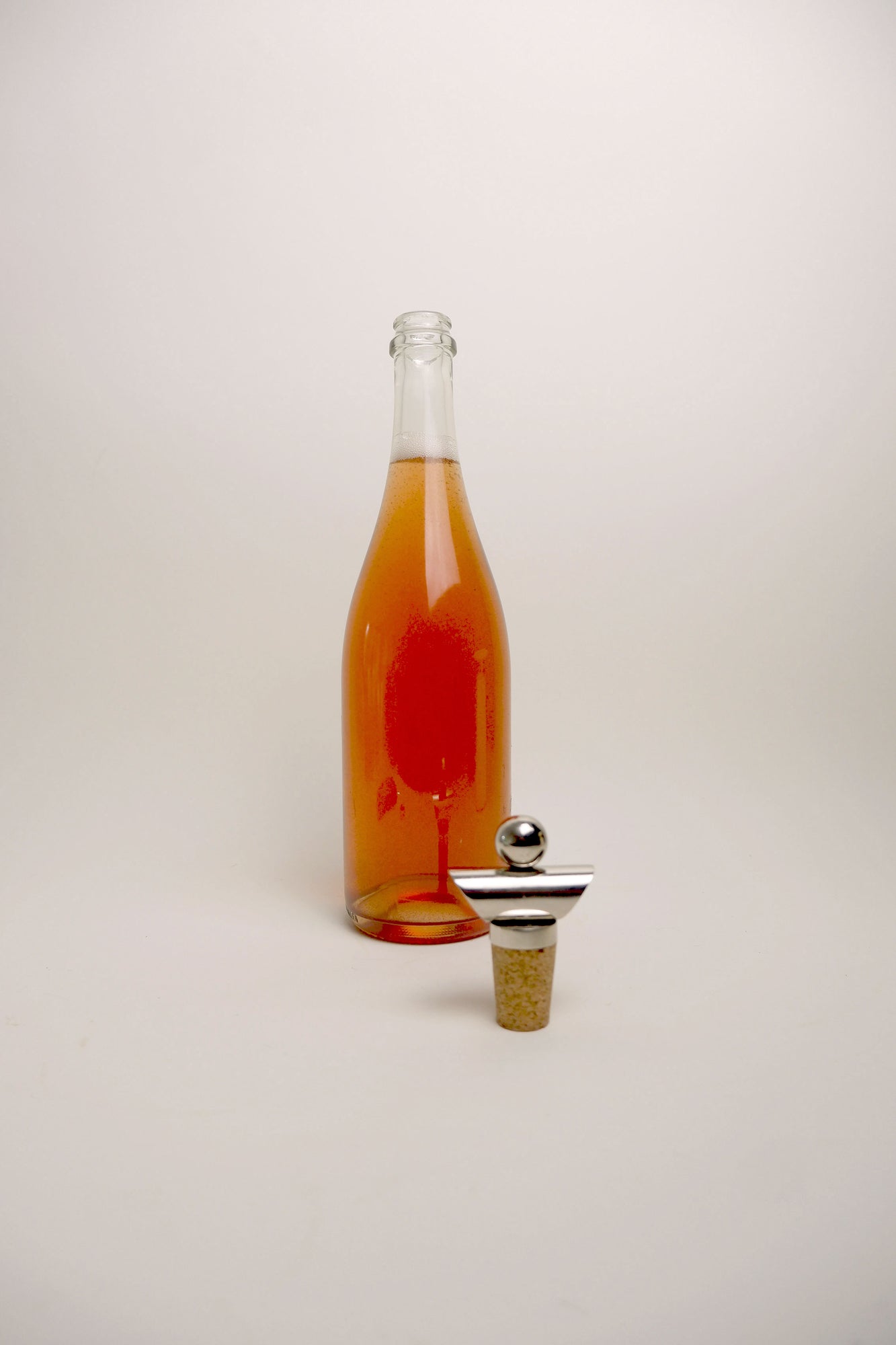 A clear glass bottle filled with amber liquid stands on a light gray studio background, with a metal-and-cork bottle stopper placed in front of it.