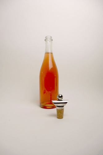A clear glass bottle filled with amber liquid stands on a light gray studio background, with a metal-and-cork bottle stopper placed in front of it.