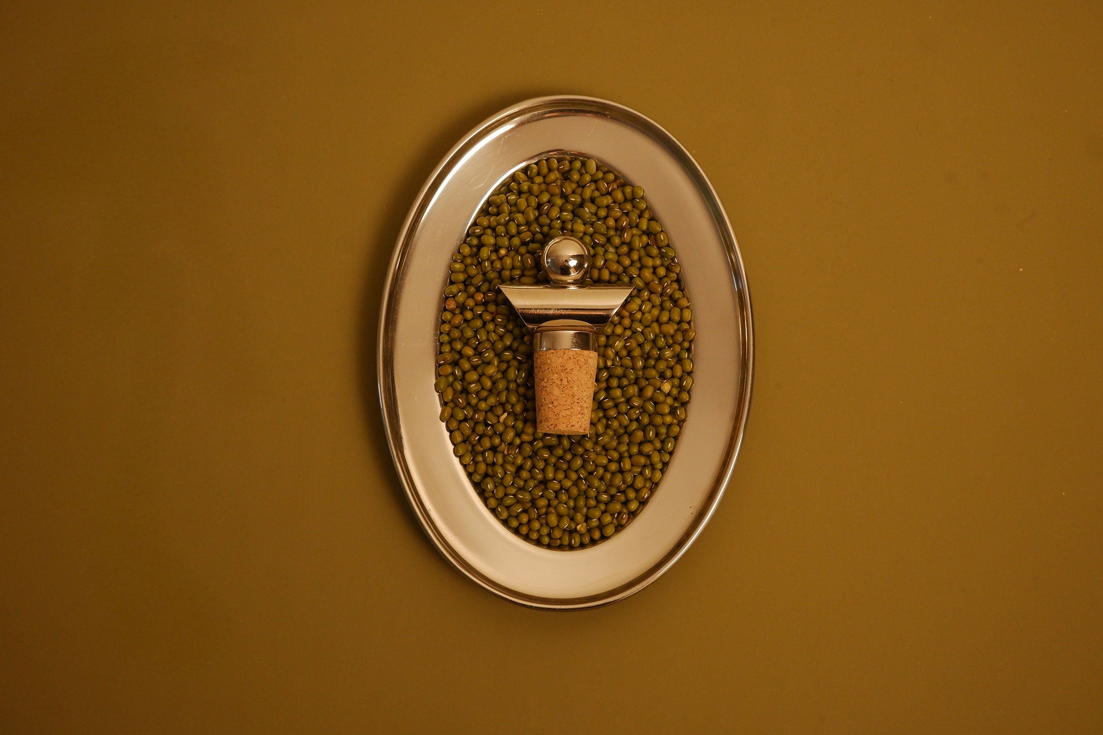 An oval wall-mounted metal holder filled with green beans, featuring a central cork stopper with a metallic cap, set against a solid mustard-yellow background.