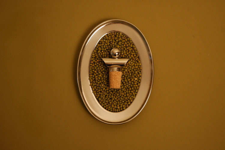 An oval wall-mounted metal holder filled with green beans, featuring a central cork stopper with a metallic cap, set against a solid mustard-yellow background.