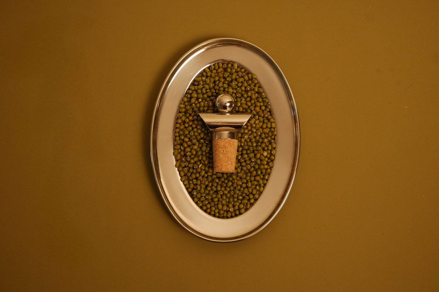 An oval wall-mounted metal holder filled with green beans, featuring a central cork stopper with a metallic cap, set against a solid mustard-yellow background.