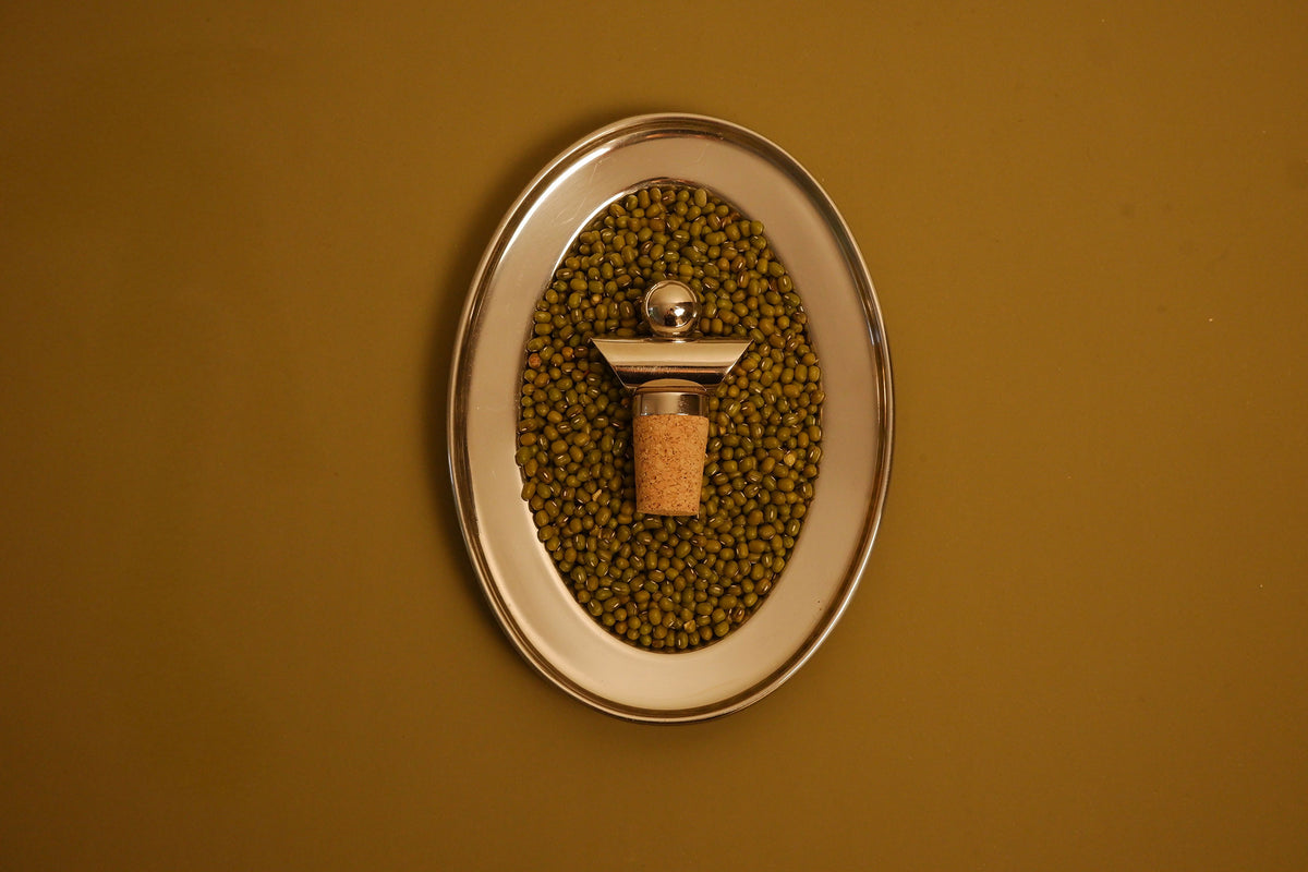 An oval wall-mounted metal holder filled with green beans, featuring a central cork stopper with a metallic cap, set against a solid mustard-yellow background.