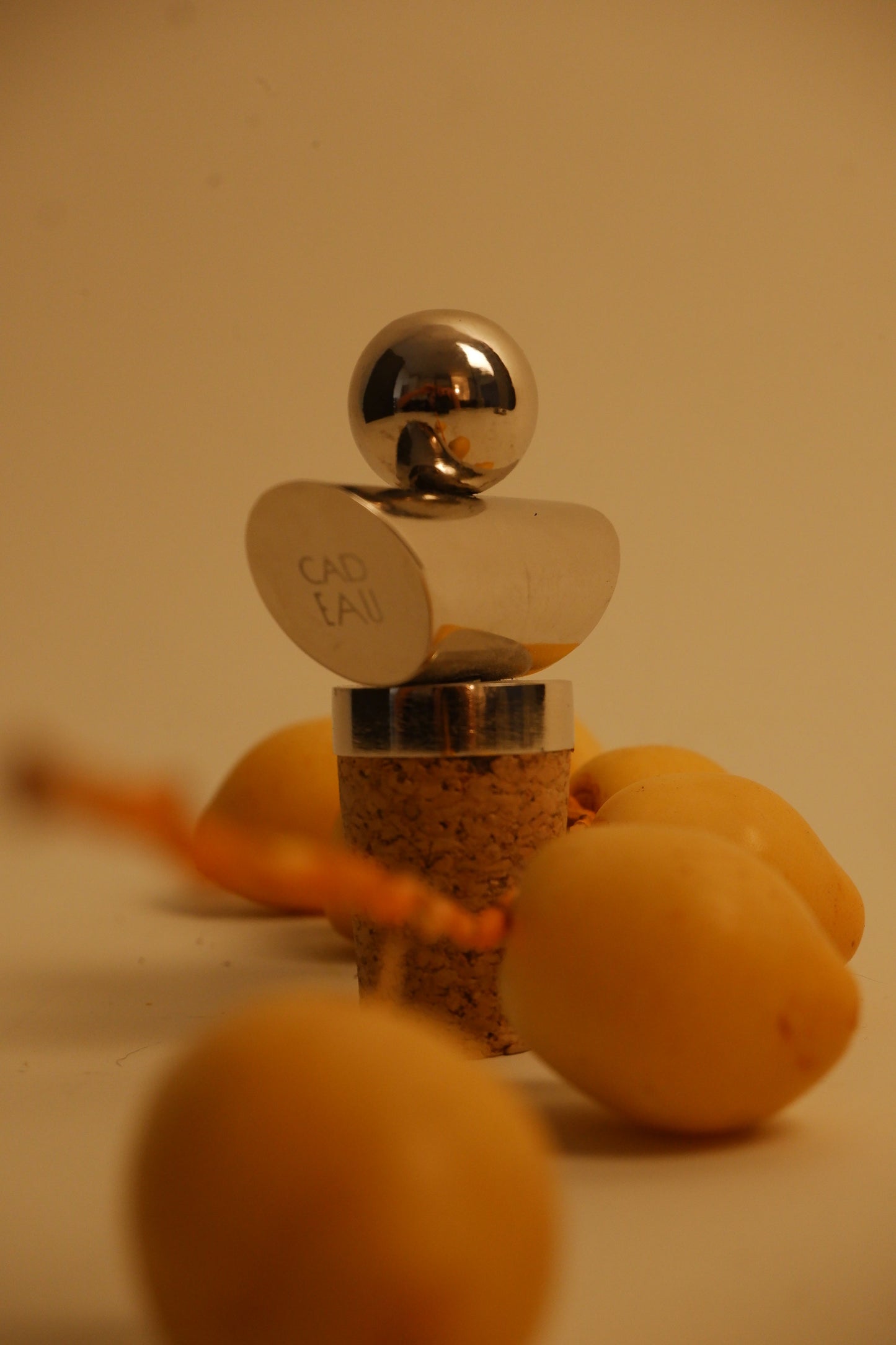 A small perfume bottle with a polished silver cap and a cork base, surrounded by several yellow-orange marigolds on a warm beige background. The cap is engraved with the text 'CAD LAU'.