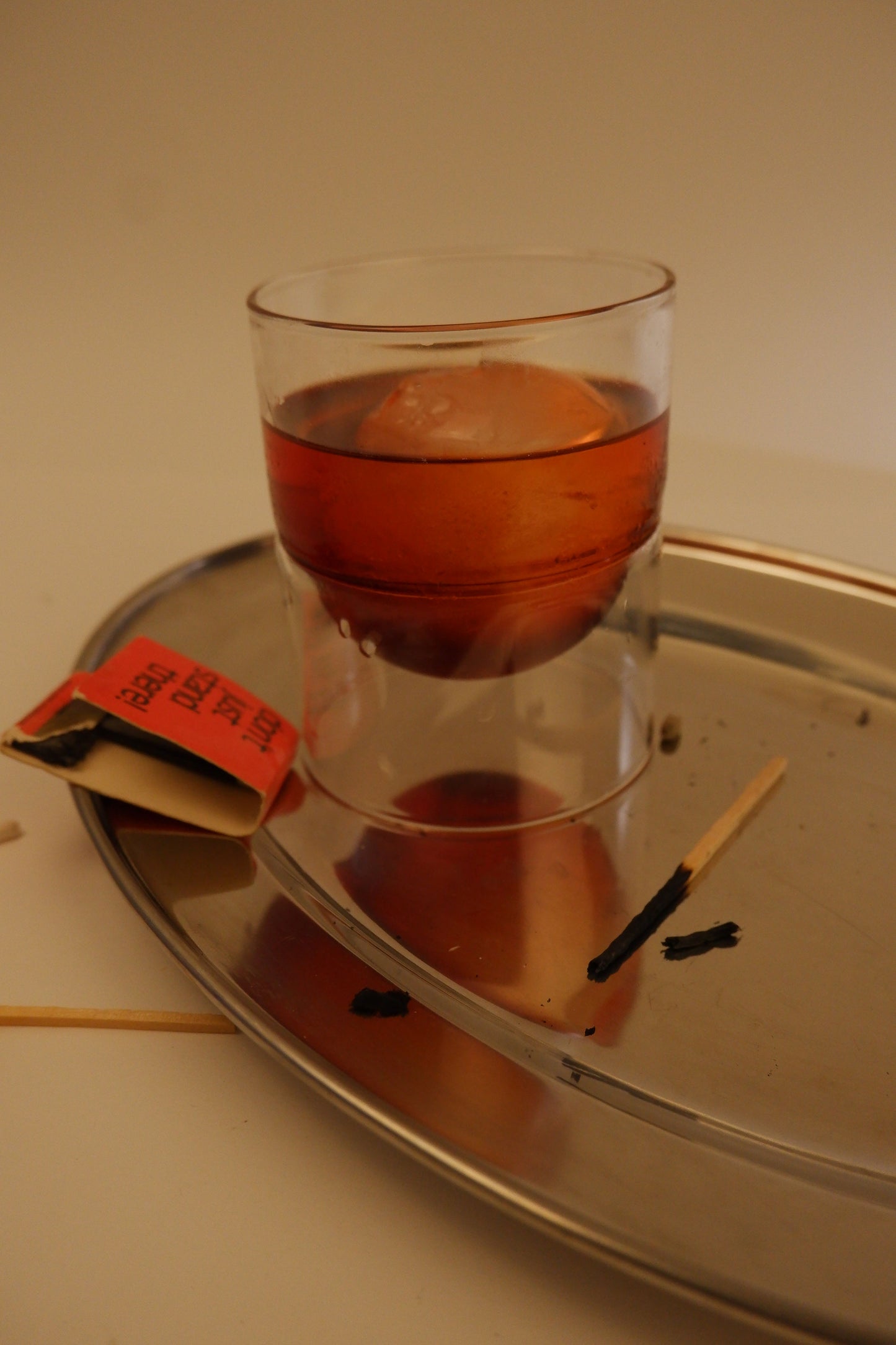 Glass of red liquid on a metal tray with a small orange object inside, on a plain background.