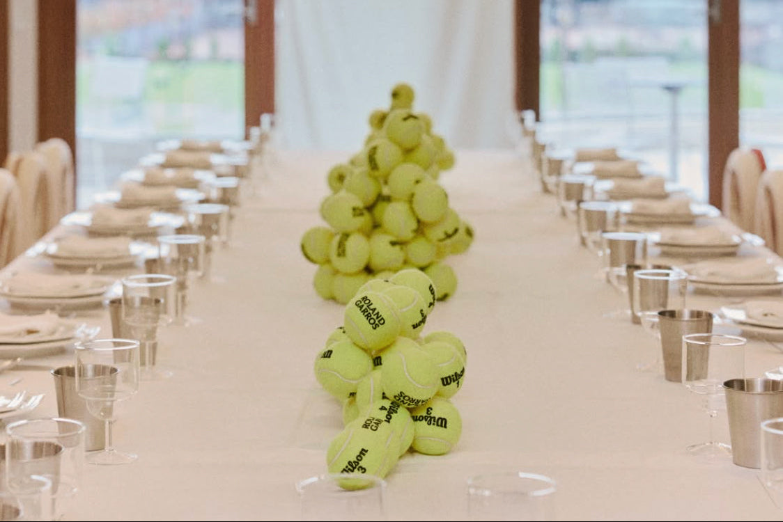 A long banquet table set with place settings and clear glasses, decorated with a central arrangement of yellow tennis balls. Large windows in the background show an outdoor view, with a white banner hanging in the center.